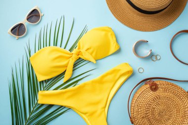 Yaz tatili konsepti. Güneş şapkalı altın yüzüklü bilezik şık güneş gözlüklü sarı bikinili rattan çanta ve izole edilmiş pastel mavi arka planda palmiye yaprakları.