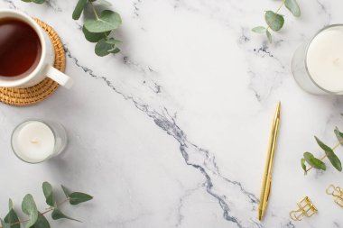 İş konsepti. En üstteki fotoğrafta, altın renkli kalem tokaları servis eden rattan çay fincanı ve beyaz mermer arka planda okaliptüs ve kopyalanmış mermer alan var.