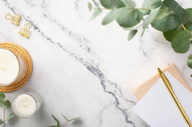 Top view photo of candles on rattan serving mat notepads gold pen binder clips and eucalyptus on white marble background with copyspace