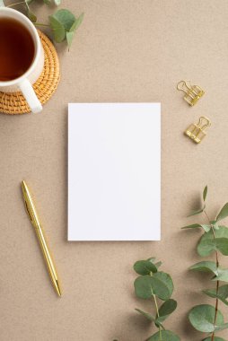 Kağıt yaprağının dikey resmi altın kalem cilt tokaları okaliptüs yaprakları ve bej arkaplan üzerinde rattan servis paspası üzerinde bir fincan çay