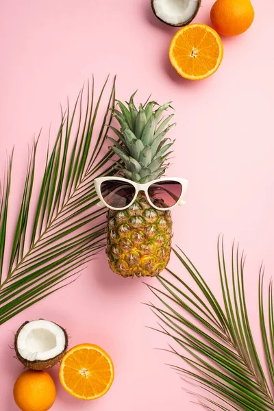Yaz havası konsepti. Güneş gözlüklü ananasın dikey fotoğrafı taze tropikal meyveler, portakallar ve izole edilmiş pastel arka planda palmiye yaprakları.