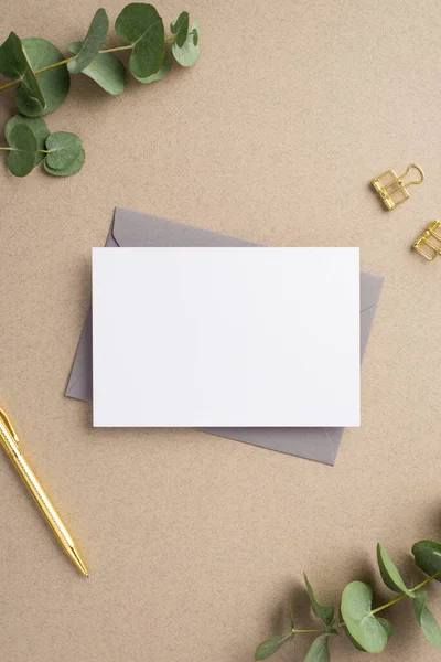 İş yeri konsepti. Kağıt yaprağının dikey resmi. Gri zarf. Altın kalem ataçları ve bej arkaplan üzerinde okaliptüs.