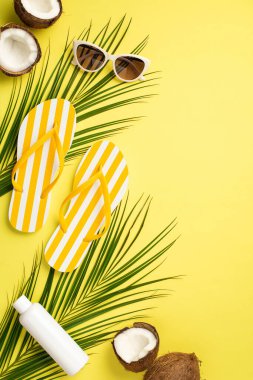 Yaz tatili konsepti. Sarı çizgili terliklerin dikey fotoğrafı güneş kremi şişesi kırılmış hindistan cevizleri ve izole edilmiş sarı arka planda palmiye yaprakları.