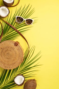 Yaz maceraları konsepti. Yuvarlak rattan çantanın dikey fotoğrafı. Güneş gözlükleri, Hindistan cevizleri ve izole edilmiş sarı arka planda palmiye yaprakları.