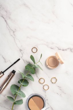 Makyaj konsepti. Beyaz mermer zemin üzerinde maskara, altın yüzükler ve okaliptüs gibi şekillendirilmiş paletin dikey görüntüsü.