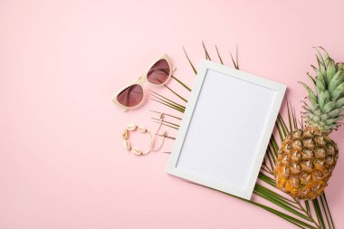 Yaz tatili kavramı. Beyaz fotoğraf çerçevesi modayı takip eden güneş gözlüklü bilezik ananas ve palmiye yapraklarının üst görüntüsü kopyalanmış pastel pembe arka plan