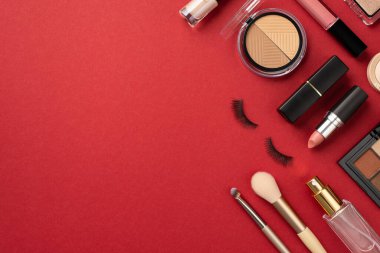 Uydurma konsept. Ruj parlatıcısının üst görüntüsü ve göz falı paletleri sahte kirpikleri makyaj fırçaları ve parfüm şişesi boş kırmızı arka planda.