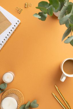 İşyeri kahve fincanının dikey fotoğrafı altın kalemler bağlayıcı mumlar rattan servis paspası ve vazo üzerinde okaliptüs ile izole edilmiş turuncu arkaplan ve ortasında kopyalanmış boşluk.