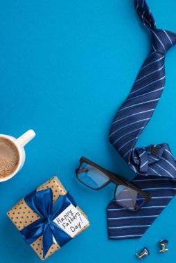 Babalar Günü konsepti. İpek fiyonklu polka nokta hediye kutusunun dikey resmi ve izole edilmiş mavi arka planda mavi kravat gözlüğü ve kol düğmeleri.