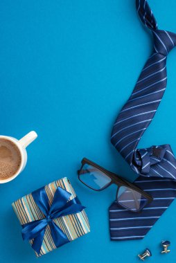Babalar Günü konsepti. İpek kurdele, mavi kravat gözlüğü, kahve fincanı ve izole edilmiş mavi arkaplanda kol düğmeleri olan şık bir hediye kutusunun dikey resmi.
