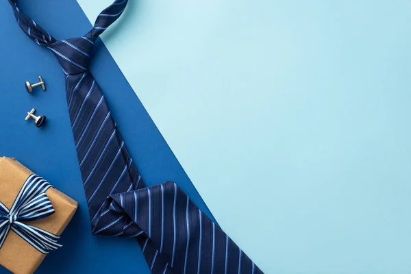 Father's Day concept. Top view photo of craft paper giftbox with ribbon bow blue striped necktie and cufflinks on bicolor blue background with empty space