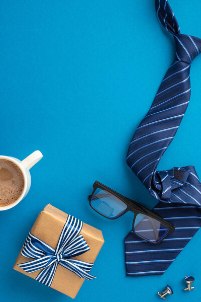 Father's Day concept. Top view vertical photo of craft paper giftbox with striped ribbon bow blue tie glasses cup of coffee and cufflinks on isolated blue background