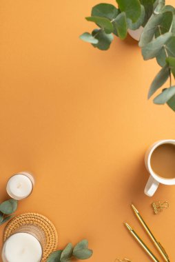 İş konsepti. Çalışma alanı kahve fincanının dikey fotoğrafı. Altın kalemler. Bağlayıcı mumlar, rattan servis paspası ve vazo üzerinde okaliptüs ve izole edilmiş turuncu arka plan.