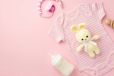 Bebek aksesuarları konsepti. Üst görüntüde pembe bebek kıyafetleri, şişe ahşap çıngırak emzik zinciri ve izole edilmiş pastel pembe arka planda örülmüş tavşan oyuncağı.