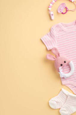Bebek konsepti. Bebek kıyafetlerinin dikey fotoğrafı pembe vücut giysisi çoraplar emzik zinciri ve izole pastel bej arka planda örülmüş tavşan çıngırağı oyuncağı.