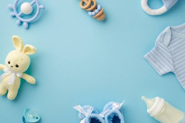 Bebek aksesuarları konsepti. Bebek kıyafetlerinin üst görüntüsü mavi gömlek örülmüş patik tavşan oyuncağı çıngırak şişesi ve izole edilmiş pastel mavi arka plan üzerinde emzik ortada boş alan