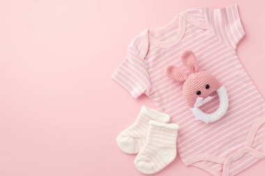 Bebek konsepti. Bebek kıyafetlerinin üst görüntüsü pembe vücut giysisi çoraplar ve örülmüş tavşan çıngırağı oyuncağı izole edilmiş pastel pembe arka planda boş alan