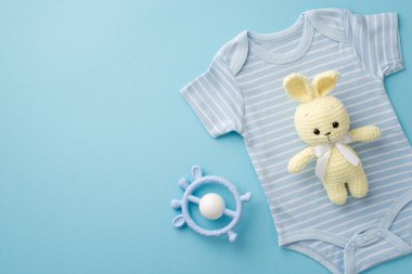 Bebek aksesuarları konsepti. Örgü ören tavşanın üst görüntüsü mavi vücut giysisi ve tenha pastel mavi arka planda çıngıraklı oyuncak