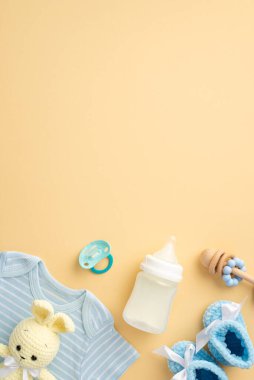 Bebek aksesuarları konsepti. Mavi tişörtün dikey fotoğrafı şişede çıngırak emziği ve izole edilmiş pastel bej arka planda örülmüş tavşan oyuncağı.