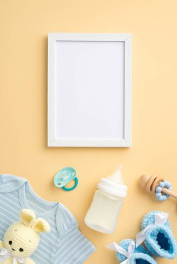 Bebek aksesuarları konsepti. En üstteki dikey fotoğraf çerçevesi mavi gömlek örgü ayakkabı şişesi ahşap çıngırak emziği ve izole edilmiş pastel bej arka planda örülmüş tavşan oyuncağı.