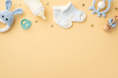 Bebek aksesuarları konsepti. Örgü örülmüş tavşanın üst görüntüsü oyuncak, mavi zincir, süt şişesi, küçük çorap emziği ve izole edilmiş pastel bej arka planda altın yıldızlar, boş alan.