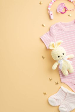Bebek aksesuarları konsepti. Bebek kıyafetlerinin dikey fotoğrafı pembe vücut giysisi çoraplar emzik zinciri örgü tavşan oyuncağı ve izole edilmiş pastel bej arka planda altın yıldızlar.