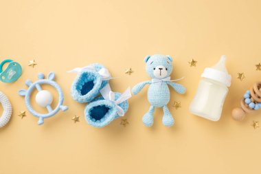 Bebek aksesuarları konsepti. Mavi örülmüş ayakların, oyuncak ayının oyuncak süt şişesinin, ahşap çıngırak emziğinin ve izole edilmiş pastel bej arka planda altın yıldızların en üst görüntüsü.