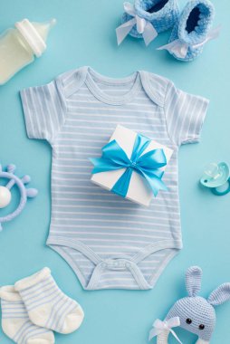 Bebek aksesuarları konsepti. Vücut giysisi çoraplarının üzerindeki hediye kutusunun dikey resmi bebek süt şişesi paçaları ve izole edilmiş pastel mavi arka planda örülmüş tavşan çıngırağı oyuncağı.