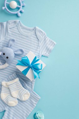 Bebek aksesuarları konsepti. Bebek kıyafetlerinin, iç çamaşırlarının, çorapların, hediye kutusunun, bağcıkların ve izole edilmiş pastel mavi arka planda örülmüş tavşan çıngıraklı oyuncakların dikey resmi.