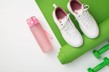 Aktif yaşam konsepti. Beyaz spor ayakkabılarının üst görüntüsü pembe su şişesi yeşil spor minderi ve izole beyaz arka planda dambıllar.