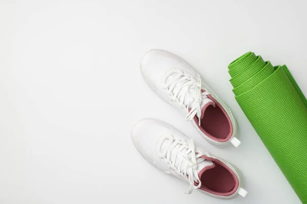 Sports accessories concept. Top view photo of white sneakers and green exercise mat on isolated white background with copyspace