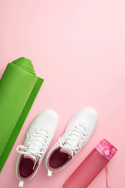 Fitness concept. Top view vertical photo of white sports footwear exercise mat and pink bottle of water on isolated pastel pink background