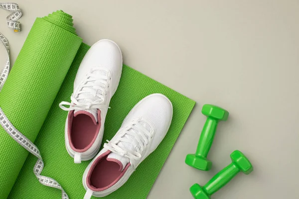 Sports accessories concept. Top view photo of dumbbells measuring tape and white sneakers over green exercise mat on isolated pastel grey background