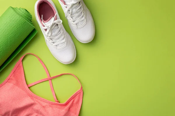 Active living concept. Top view photo of white sneakers pink sports bra and green exercise mat on isolated green background