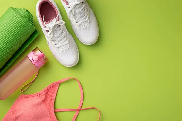 Fitness concept. Top view photo of white sneakers pink sports top bottle of water and green exercise mat on isolated green background