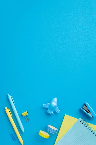 Back to school concept. Top view vertical photo of yellow and blue stationery notebooks pens plane shaped sharpener binder clip adhesive tape and stapler on isolated blue background with copyspace