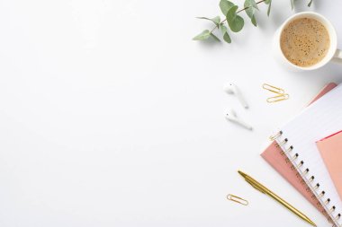 Business concept. Top view photo of workplace stack of pink planners gold pen clips eucalyptus cup of coffee and wireless earbuds on isolated white background with copyspace