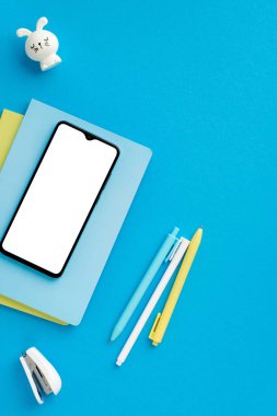 Okul konseptine geri dönelim. Üst görünümde akıllı telefonun dikey fotoğrafı defter yığınının üzerinde dolgu kalemler zımba ve tavşan şekilli bileyiciler izole edilmiş mavi arka planda boş alanla