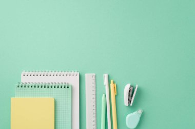 Okul aksesuarları konsepti. Kırtasiye defterlerinin en üst görünüm fotoğrafı boş arkaplan ile izole edilmiş pastel yeşil arkaplan üzerinde kalem yuvarlak düzeltme bant cetvel ve mini zımba