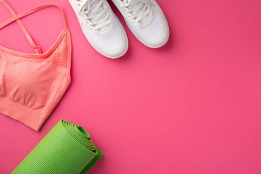 Fitness accessories concept. Top view photo of green yoga mat pink sports top and white shoes on isolated pink background with copyspace