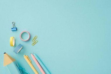 Okul konseptine geri dönelim. Okul malzemelerinin en üst görüntüsü renkli kalemler mavi kalem kutusu yapıştırıcı bant ve izole edilmiş pastel mavi arkaplan üzerine yapıştırıcı klipleri