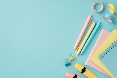 Okul konseptine geri dönelim. Okul malzemelerinin en üst görüntüsü not defterleri, yapıştırıcı bant klipleri ve izole edilmiş pastel mavi arka plan üzerindeki işaretleyici kalemler.