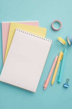 Okul malzemeleri konsepti. Renkli not defteri yığınının dikey üst görünümü. İzole edilmiş pastel mavi arkaplan üzerinde kalem klasörü ve yapıştırıcı bant boş alan.