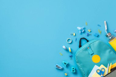 Not defterleri ve makaslı sırt çantasının en üst görüntüsü. Plastik alfabe harfi, klasör, uçak şekilli silgi, yapıştırıcı, yapıştırıcı, bant zımbası, boş alan ile mavi arkaplanı izole ediyor.