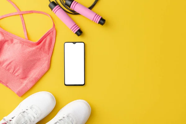 Fitness accessories concept. Top view photo of pink sports top white sneakers skipping rope and smartphone on isolated yellow background with copyspace