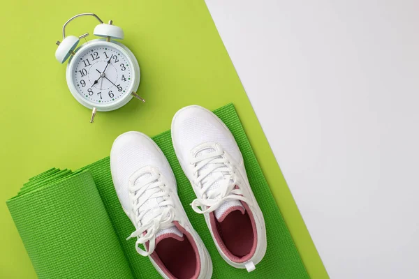 Sports concept. Top view photo of white sneakers over green exercise mat and alarm clock on bicolor green and white background with copyspace