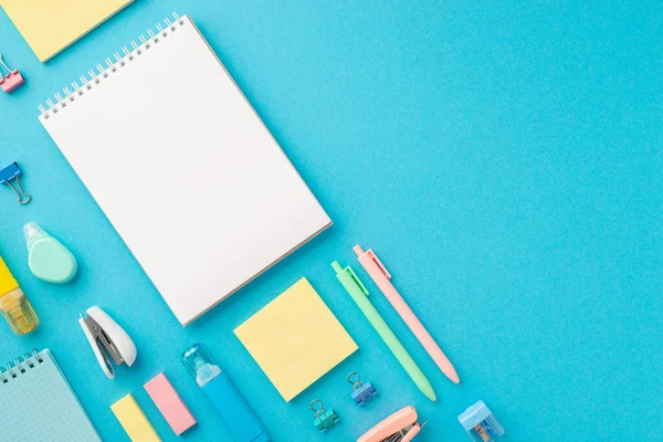 Back to school concept. Top view photo of ordered stationery open spiral notepad sharpener binder clips correction pens eraser stapler paper sticky notes on isolated blue background with blank space