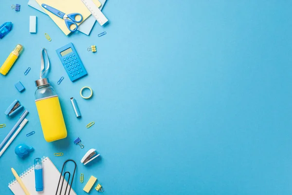 Back to school concept. Top view photo of school supplies yellow drink bottle correction pen clip stapler eraser notepad adhesive tape calculator scissors ruler isolated blue background with copyspace