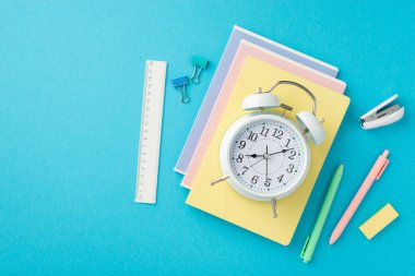 Okul konseptine geri dönelim. Alarm saatinin üst görünümü, not defterleri zımba telleri silgi cetvelleri ve izole edilmiş mavi arkaplan üzerindeki klasör klipleri üzerinedir.