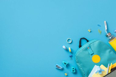 Mavi okul çantasının, not defterleriyle ve uçak şeklindeki kalemlerle en iyi fotoğrafı. Sivri uçlu içki şişesi, plastik alfabe harfleri. Bağlayıcı raptiyeler, izole edilmiş mavi arka planda yapıştırıcı.